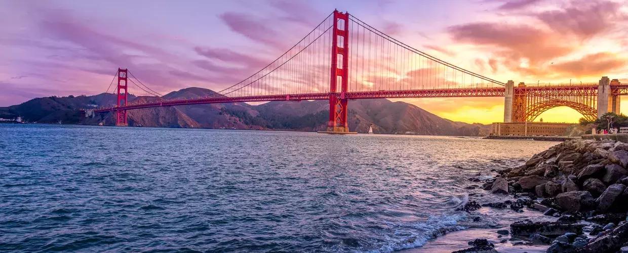 Le 金门大桥 au coucher du soleil avec un ciel multicolore et la baie de San Francisco au premier plan.
